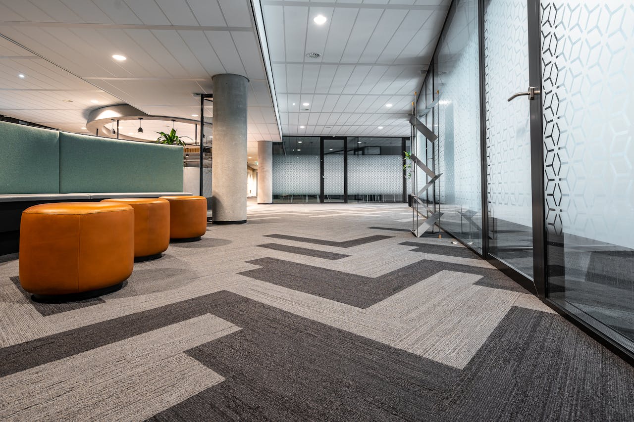 who-we-are Spacious and modern office corridor featuring glass doors, geometric carpet patterns, and leather seating.